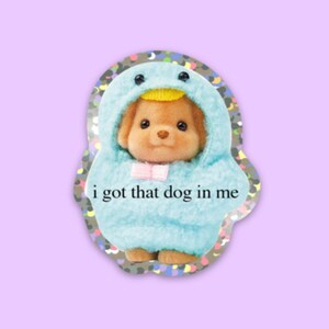May include: A sticker featuring a small, brown dog wearing a blue and yellow penguin costume. The sticker has a glittery background and the text "i got that dog in me" printed on it.