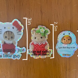 May include: Three holographic stickers featuring cartoon animals wearing costumes. The first sticker is 3 inches tall and features a cat wearing a Hello Kitty costume with the text "i am simply just a little guy". The second sticker is 2.5 inches tall and features a cat wearing a green and white dress with the text "i can act rather silly at times". The third sticker is 2 inches tall and features a dog wearing a blue penguin costume with the text "i got that dog in me".