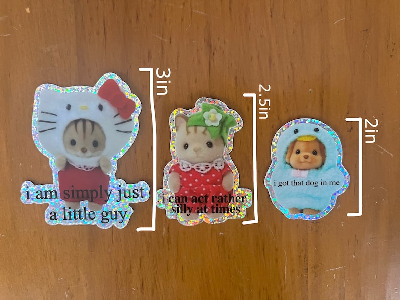 May include: Three holographic stickers featuring cartoon animals wearing costumes. The first sticker is 3 inches tall and features a cat wearing a Hello Kitty costume with the text "i am simply just a little guy". The second sticker is 2.5 inches tall and features a cat wearing a green and white dress with the text "i can act rather silly at times". The third sticker is 2 inches tall and features a dog wearing a blue penguin costume with the text "i got that dog in me".