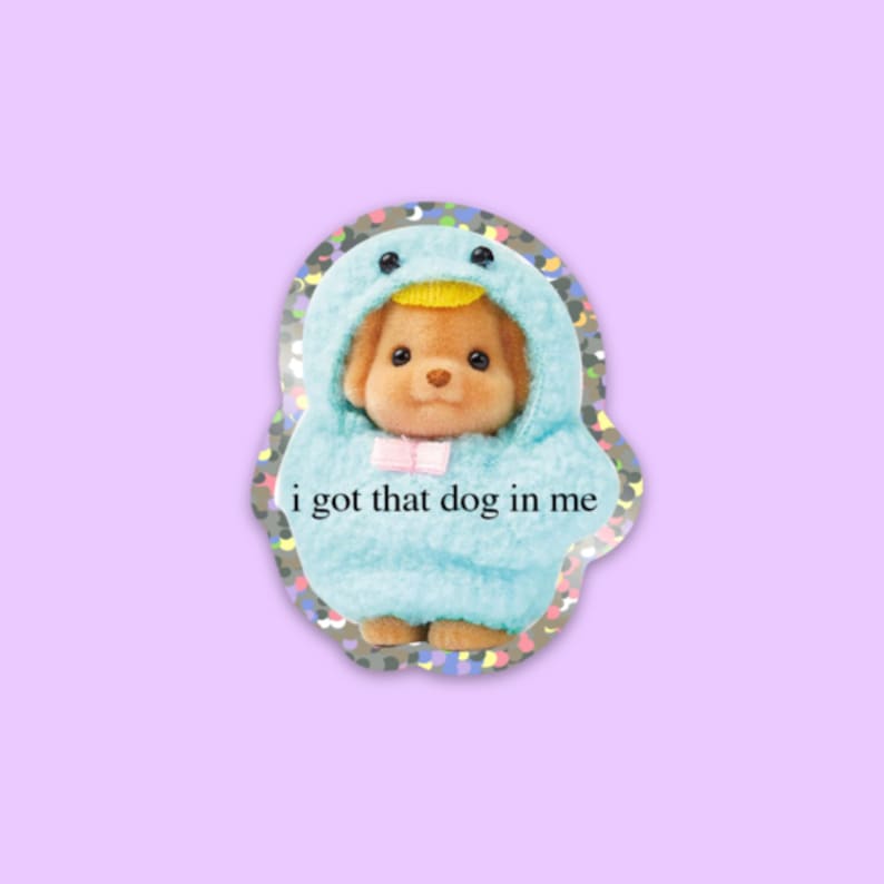 May include: A sticker featuring a small, brown dog wearing a blue and yellow penguin costume. The sticker has a glittery background and the text "i got that dog in me" printed on it.