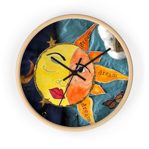 Puede incluir: Un reloj de pared redondo con marco de madera presenta un diseño caprichoso de sol y luna. El sol es amarillo y naranja con una cara sonriente y un ojo cerrado, mientras que la luna es amarilla con un ojo guiñando y labios rojos. La esfera del reloj está rodeada de un fondo azul y verde con estrellas y la palabra "dream" repetida tres veces.
