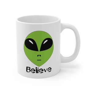 Puede incluir: Taza de café de cerámica blanca con una cara de extraterrestre verde y la palabra "Believe" impresa en negro.