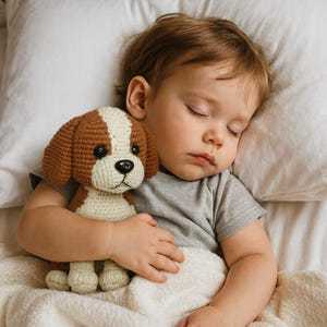 May include: A sleeping child cuddles a handmade crochet dog toy. The toy features brown and white sections, with black button eyes and a black nose. The child is wearing a gray t-shirt and is nestled in a white bed.