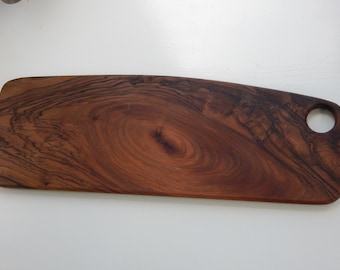 Könnte beinhalten: Ein rechteckiges Holzschneidebrett mit dunkelbraunem Finish und natürlichem Holzmaserungsmuster. Das Brett hat einen abgerundeten Griff mit einem kreisförmigen Loch. Das Brett ist etwa 46 cm lang.