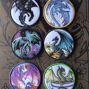 Peut inclure: Ensemble de six écussons brodés circulaires représentant des dragons. Chaque écusson présente un dragon détaillé sur fond blanc, avec des couleurs variées comme le noir, le bleu, le violet et le vert. Les écussons ont une bordure cousue et sont posés sur un fond à motifs.