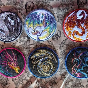 May include: Six embroidered patches featuring dragon designs. The patches are circular with various color schemes, including a yin-yang dragon, a dragon with blue and green wings, and a fiery orange dragon. The patches are on a patterned background.