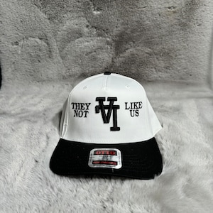 Puede incluir: Gorra de béisbol blanca con visera negra. La parte delantera de la gorra tiene bordado en negro el texto "THEY NOT LIKE US" con una "V" estilizada en el medio. La parte posterior de la gorra tiene una etiqueta negra con texto blanco que dice "OTTO CLASSIC FIT CROWN MID PROFILE MAXIMUM COMFORT".