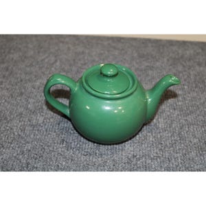 May include: A solid green ceramic teapot with a rounded body, a curved handle, a short spout, and a matching removable lid with a small knob handle on top. The teapot features a smooth, glossy finish and a simple, functional design.