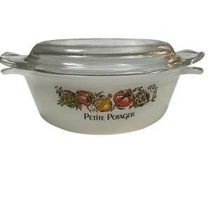 May include: A white, vintage casserole dish with a clear glass lid. The dish features a colorful vegetable design with the words "Petite Potager" printed below. The dish has two handles and is suitable for cooking and serving.