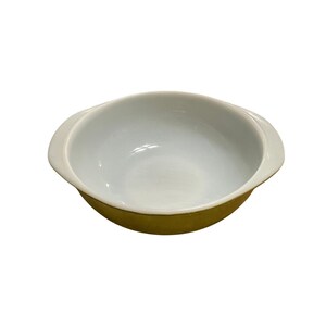 May include: A round ceramic baking dish featuring a smooth white interior and a solid mustard yellow exterior. The dish includes two integrated side handles for gripping, with a shallow, wide basin design suitable for various culinary preparations.