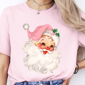 May include: A light pink t-shirt featuring a vintage Santa Claus graphic. The Santa Claus illustration has a pink hat, a white beard, and a smiling face. The shirt is paired with blue jeans.