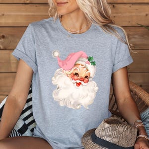 May include: A light gray t-shirt featuring a vintage Santa Claus graphic. The Santa has a pink hat, rosy cheeks, and a long white beard. The shirt is short-sleeved and has rolled-up sleeves.