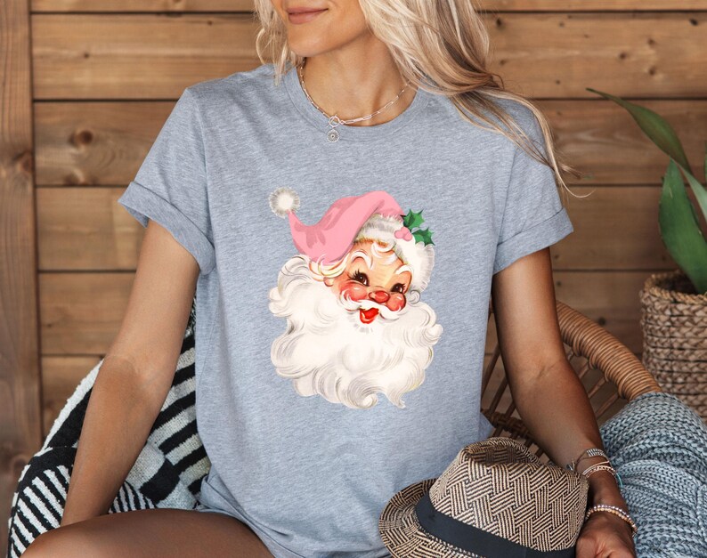 May include: A light gray t-shirt featuring a vintage Santa Claus graphic. The Santa has a pink hat, rosy cheeks, and a long white beard. The shirt is short-sleeved and has rolled-up sleeves.