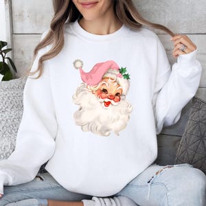 May include: White sweatshirt featuring a vintage Santa Claus graphic. The Santa has a pink hat, rosy cheeks, and a long white beard. The design includes holly leaves and a pom-pom on the hat, perfect for the holiday season.