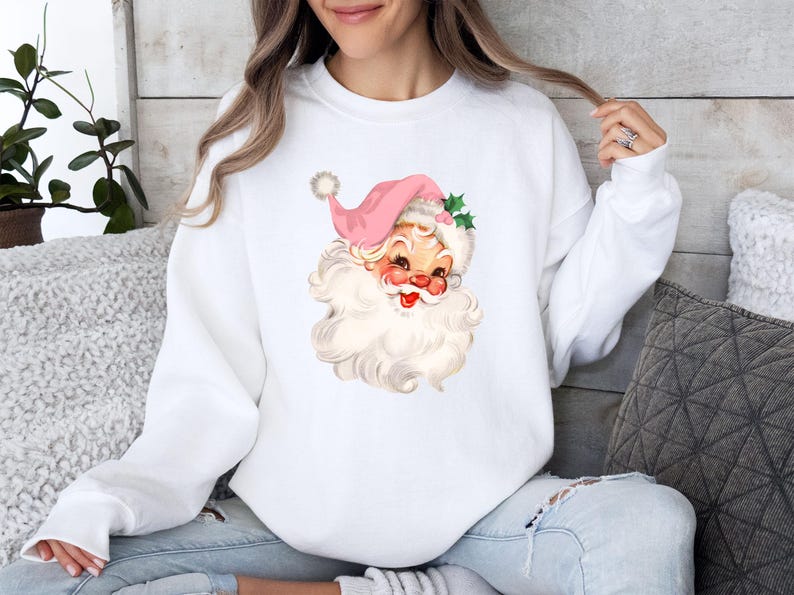 May include: White sweatshirt featuring a vintage Santa Claus graphic. The Santa has a pink hat, rosy cheeks, and a long white beard. The design includes holly leaves and a pom-pom on the hat, perfect for the holiday season.