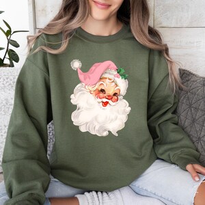 May include: An olive green sweatshirt featuring a vintage Santa Claus graphic. The Santa has a pink hat, rosy cheeks, and a large white beard. The sweatshirt is a comfortable, casual style.