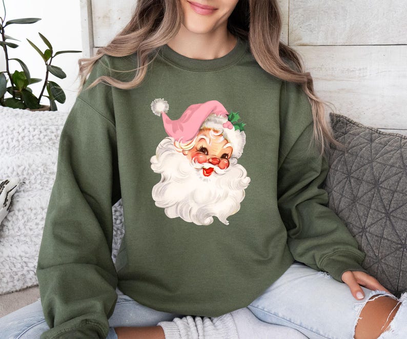 May include: An olive green sweatshirt featuring a vintage Santa Claus graphic. The Santa has a pink hat, rosy cheeks, and a large white beard. The sweatshirt is a comfortable, casual style.