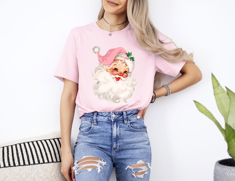 May include: A light pink t-shirt featuring a vintage Santa Claus graphic. The Santa Claus illustration has a pink hat, a white beard, and a smiling face. The shirt is paired with blue jeans.
