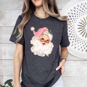 May include: Dark gray t-shirt featuring a vintage Santa Claus graphic. The Santa illustration has a pink hat, a white beard, and rosy cheeks. The design is a festive, retro-style Christmas theme.