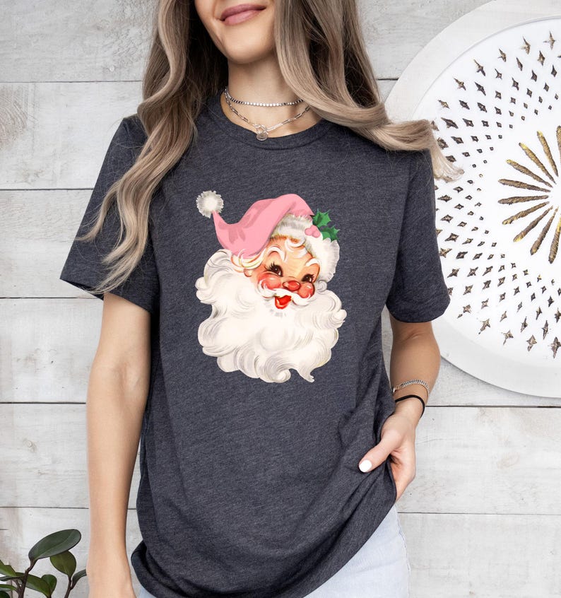May include: Dark gray t-shirt featuring a vintage Santa Claus graphic. The Santa illustration has a pink hat, a white beard, and rosy cheeks. The design is a festive, retro-style Christmas theme.