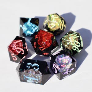 May include: Seven polyhedral dice with black resin exteriors and colorful, glittery interiors. The dice feature red, pink, blue, and green accents, with numbers in various colors. The dice are arranged in a circular pattern on a white surface.
