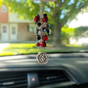 May include: A silver car charm with a sun and moon design hangs from a rearview mirror. The charm is attached to a string of red, black, and clear beads.
