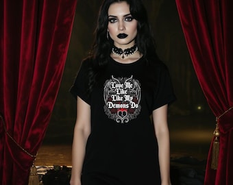 Op de afbeelding: Zwart T-shirt met de tekst "Love Me Like My Demons Do" in een gotische stijl. Het ontwerp heeft een hart- en vleugelmotief. Het model draagt een zwarte choker en visnetkousen.
