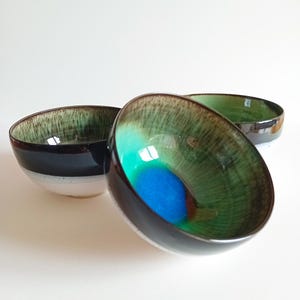 Peut inclure: Trois bols en céramique avec une finition brillante. Les bols ont un extérieur foncé, une bande blanche et un intérieur vert et bleu. Les bols sont disposés sur une surface blanche. Les bols sont probablement destinés à servir des aliments.
