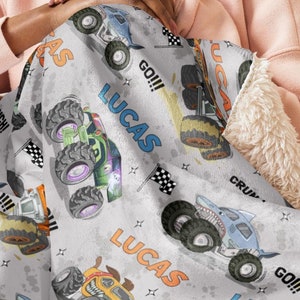 Op de afbeelding: Een grijs en wit fleece deken met een herhalend patroon van monstertrucks. De trucks zijn blauw, groen, oranje en geel. Het deken heeft de naam "Lucas" meerdere keren erop gedrukt. Het deken wordt vastgehouden door een persoon die op een bank zit.