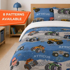 May include: A blue and gray monster truck themed bedding set. The set includes a comforter, pillowcases, and a framed picture of a monster truck. The comforter features various colorful monster trucks on a blue and gray background. The text "ATTICUS" is also visible.