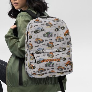 May include: A gray backpack with a white background and a pattern of monster trucks. The backpack has a black zipper and a black adjustable strap. The name "JORDAN" is printed in orange on the front of the backpack.