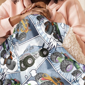Puede incluir: Una manta de franela azul y blanca con un estampado de estilo c&oacute;mic de monster trucks. Los camiones son de varios colores, incluyendo naranja, verde y azul.