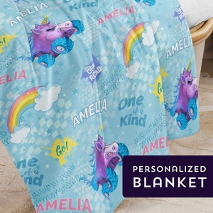May include: A blue personalised blanket with a unicorn design. The blanket features rainbows, stars, and the text "Amelia" and "One of a Kind".