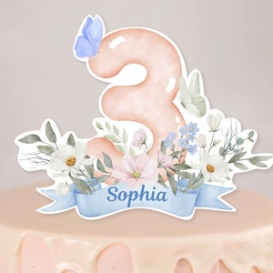 Op de afbeelding: Een roze nummer drie taarttopper met een blauwe vlinder, witte en roze bloemen en groene bladeren. De topper heeft een blauw lint met de naam "Sophia" erop.