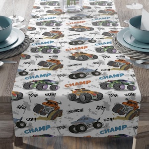 May include: White table runner with a repeating pattern of cartoon monster trucks. The trucks are orange, green, and blue with black wheels. The text "CHAMP" is repeated throughout the pattern, along with the words "GO!!!", "WOW!" and "CRUNCH!"