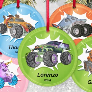 May include: Set of six round Christmas ornaments with monster truck designs. Each ornament features a different monster truck illustration in vibrant colors, with names and the year 2024 or 2020 printed below the image. Red ribbons are attached for hanging.