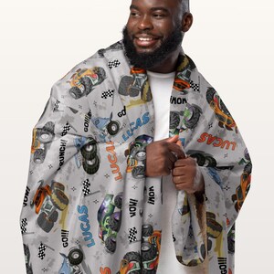 Op de afbeelding: Een grijze fleece deken met een herhalend patroon van monstertrucks, vlaggen en de naam "Lucas" in oranje, blauw en groen. De deken wordt vastgehouden door een persoon.