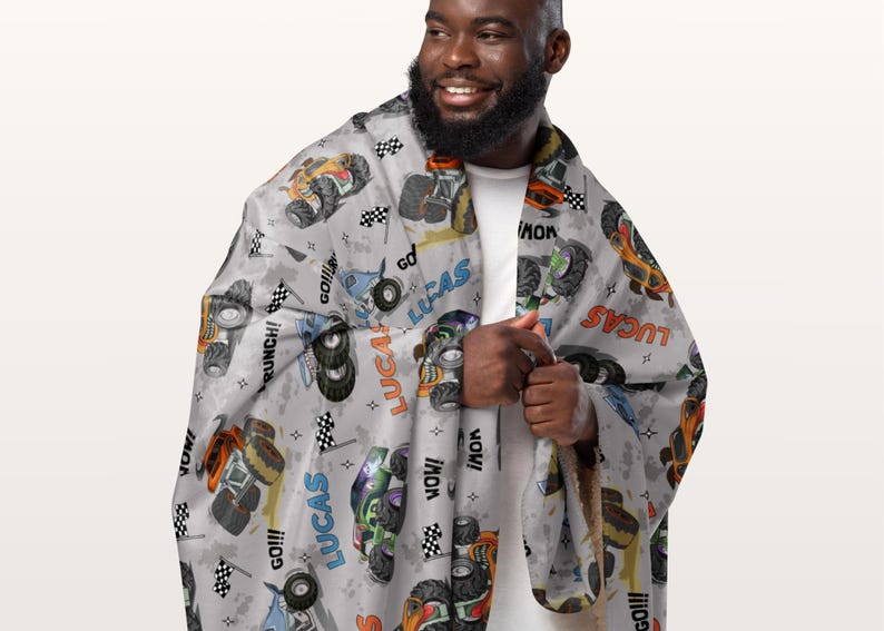 Op de afbeelding: Een grijze fleece deken met een herhalend patroon van monstertrucks, vlaggen en de naam "Lucas" in oranje, blauw en groen. De deken wordt vastgehouden door een persoon.