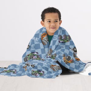 May include: A blue and white checkered fleece blanket with a monster truck design. The blanket features a variety of monster trucks in different colors and designs. The blanket is wrapped around a child.