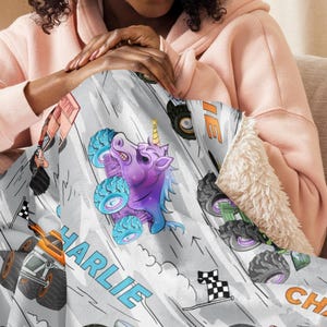 May include: A light grey blanket with a monster truck design. It features a purple unicorn monster truck, racing flags, and the name "CHARLIE" in blue and orange. The blanket has a soft, white sherpa lining for added comfort.