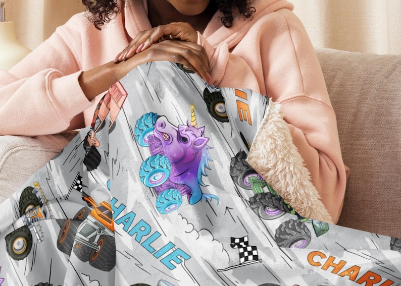 May include: A light grey blanket with a monster truck design. It features a purple unicorn monster truck, racing flags, and the name "CHARLIE" in blue and orange. The blanket has a soft, white sherpa lining for added comfort.