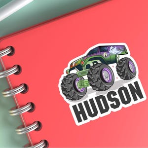 May include: A sticker featuring a cartoon monster truck with large tires, green and purple accents, and a ghost graphic. The name "HUDSON" is printed in bold, white letters below the truck.