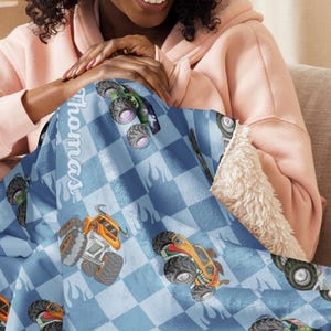 May include: A blue and white checkered fleece blanket with a monster truck design. The blanket features a personalized name, "Thomas", in white script.