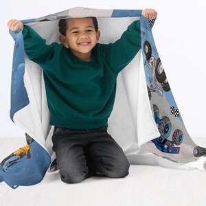 May include: A child is holding up a blue and white blanket with a monster truck design. The blanket features illustrations of monster trucks, checkered flags, and other racing elements. The child is wearing a green sweater and dark jeans.