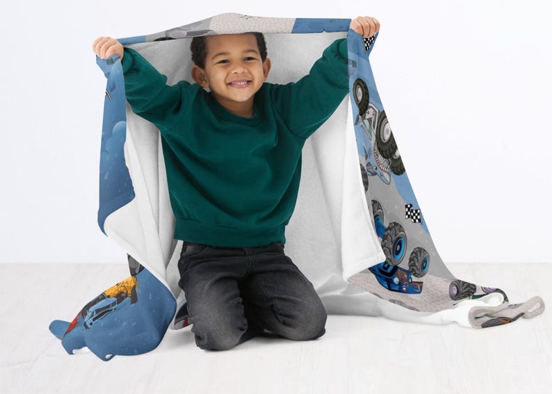 May include: A child is holding up a blue and white blanket with a monster truck design. The blanket features illustrations of monster trucks, checkered flags, and other racing elements. The child is wearing a green sweater and dark jeans.