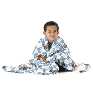 May include: A blue and white chequered fleece blanket with a monster truck design and the name "Henry" printed on it. A child is sitting on the floor wrapped in the blanket.