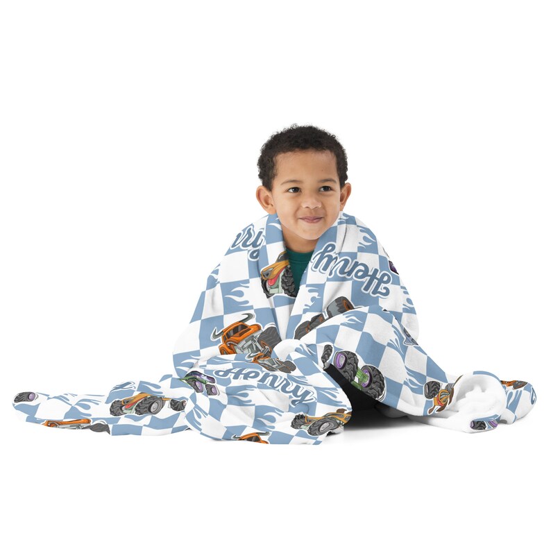 May include: A blue and white chequered fleece blanket with a monster truck design and the name "Henry" printed on it. A child is sitting on the floor wrapped in the blanket.