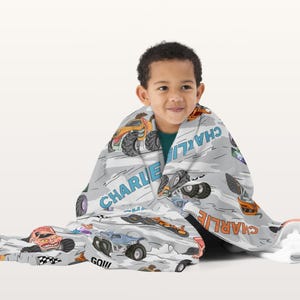 May include: A light grey blanket with a repeating pattern of colourful monster trucks. The name "CHARLIE" is printed in blue and orange. The blanket is wrapped around a child.