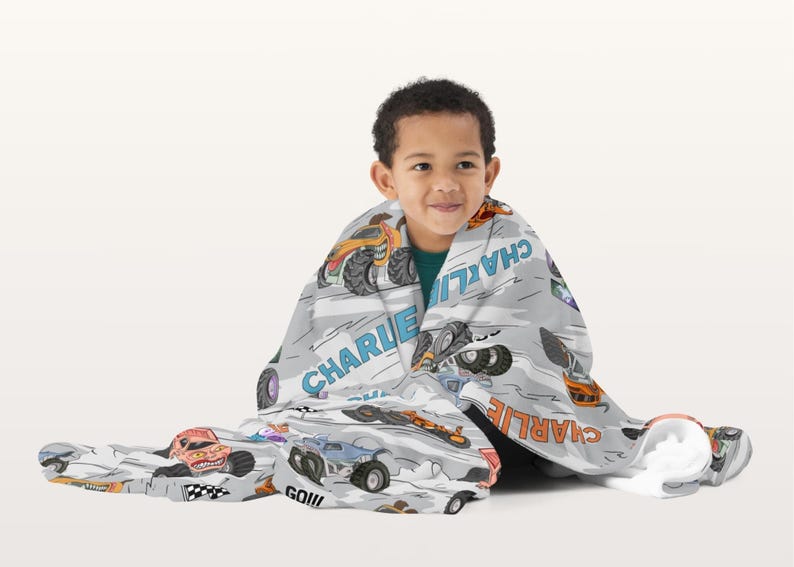 May include: A light grey blanket with a repeating pattern of colourful monster trucks. The name "CHARLIE" is printed in blue and orange. The blanket is wrapped around a child.