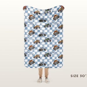 May include: A blue and white chequered baby blanket with a repeated pattern of monster trucks. The trucks are orange, black, and grey. The blanket has the name "Henry" printed on each truck. The blanket is 76 cm by 102 cm.